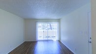 This single-floor space offers a clean, functional layout with a bright and open living area as its centerpiece. The living area is finished with rich wood-style hardwood flooring that reflects natural light beautifully, creating a warm and inviting atmosphere. A full-width sliding glass door serves as a prominent architectural feature, allowing abundant daylight to flood the interior while providing direct views of the surrounding outdoor environment. Sheer vertical blinds frame the sliding door, offering privacy control without compromising the sense of openness. The walls are finished in a neutral, light tone that complements the flooring and enhances the airy feel of the room. A textured popcorn ceiling adds a classic residential character to the space. The overall design is straightforward and versatile, making it well-suited for a variety of furnishing arrangements. The seamless connection between the interior and the outdoors through the sliding glass door is a standout element, contributing to the space's bright, open ambiance throughout the day.