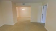 This single-floor space presents a clean, open layout with interconnected living areas that flow naturally into one another. The interior is finished with warm beige carpeting throughout, providing a cohesive and comfortable feel underfoot. Walls are painted in a soft, neutral cream tone, contributing to a bright and airy atmosphere.

A defining architectural feature is the wide open passageway connecting the main living area to an adjacent room, likely a dining space, framed by a squared archway that adds a sense of depth and dimension. The dining area is highlighted by a classic multi-bulb chandelier suspended from the ceiling, serving as a focal point and adding a touch of traditional elegance.

Ceiling-mounted HVAC vents are integrated discreetly into the layout, ensuring functional climate control without disrupting the overall aesthetic. White baseboards line the perimeter of both rooms, providing a clean contrast against the carpeted floors. A door visible along one wall suggests access to an additional room or closet. Overall, the space offers a neutral, move-in-ready canvas well-suited for a variety of interior styles.