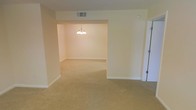 This single-floor space presents a clean, open layout with interconnected living areas that flow naturally into one another. The interior is finished with warm beige carpeting throughout, providing a cohesive and comfortable feel underfoot. Walls are painted in a soft, neutral cream tone, contributing to a bright and airy atmosphere.

A defining architectural feature is the wide open passageway connecting the main living area to an adjacent room, likely a dining space, framed by a squared archway that adds a sense of depth and dimension. The dining area is highlighted by a classic multi-bulb chandelier suspended from the ceiling, serving as a focal point and adding a touch of traditional elegance.

Ceiling-mounted HVAC vents are integrated discreetly into the layout, ensuring functional climate control without disrupting the overall aesthetic. White baseboards line the perimeter of both rooms, providing a clean contrast against the carpeted floors. A door visible along one wall suggests access to an additional room or closet. Overall, the space offers a neutral, move-in-ready canvas well-suited for a variety of interior styles.