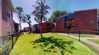 This residential community presents a classic and functional design characterized by sturdy red brick exteriors accented with navy blue and gray siding, creating a distinctive and cohesive aesthetic throughout the property. The grounds are well-maintained, featuring generous stretches of lush green lawn shaded by mature trees, providing a welcoming outdoor environment for residents. Walkways and fenced pathways connect the buildings, with metal staircases offering access between levels of the multi-unit structures. The community's standout feature is its outdoor amenity area, which includes a sparkling swimming pool surrounded by a spacious stone-tiled patio. A pergola provides partial shade over an outdoor seating and dining area, while a built-in outdoor kitchen with a grill adds convenience for entertaining. Large windows in the clubhouse or common building overlook the pool area, enhancing the connection between indoor and outdoor spaces. The entry to this central building features a glass-paneled door framed by exposed brick and dark wood siding, lending a modern touch to the traditional architecture. The patio's multicolored stone tile flooring adds texture and visual interest to the overall outdoor space, making it an inviting area for relaxation and social gatherings.