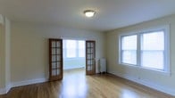 This single-floor space showcases a thoughtfully designed interior that blends classic architectural details with a clean, functional layout. The living area is highlighted by warm hardwood floors that run throughout, reflecting natural light and lending a sense of warmth and continuity to the space. A standout feature is the set of classic wood-framed French doors with divided-light glass panes, elegantly separating two interconnected rooms while maintaining an open, airy flow between them. Multiple windows fitted with blinds allow generous amounts of natural light to fill each room, creating a bright and welcoming atmosphere. A traditional cast-iron radiator adds a charming, period-appropriate touch, hinting at the space's classic character. The walls are finished in neutral tones, providing a versatile backdrop suited to a variety of interior styles. Flush-mount ceiling lighting provides functional illumination, complementing the abundant daylight. The overall ambiance is one of understated elegance — clean lines, natural materials, and timeless architectural elements come together to create a space that is both practical and full of character.