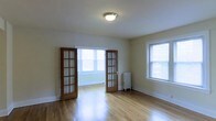 This single-floor space showcases a thoughtfully designed interior that blends classic architectural details with a clean, functional layout. The living area is highlighted by warm hardwood floors that run throughout, reflecting natural light and lending a sense of warmth and continuity to the space. A standout feature is the set of classic wood-framed French doors with divided-light glass panes, elegantly separating two interconnected rooms while maintaining an open, airy flow between them. Multiple windows fitted with blinds allow generous amounts of natural light to fill each room, creating a bright and welcoming atmosphere. A traditional cast-iron radiator adds a charming, period-appropriate touch, hinting at the space's classic character. The walls are finished in neutral tones, providing a versatile backdrop suited to a variety of interior styles. Flush-mount ceiling lighting provides functional illumination, complementing the abundant daylight. The overall ambiance is one of understated elegance — clean lines, natural materials, and timeless architectural elements come together to create a space that is both practical and full of character.