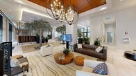 This expansive, single-floor lobby is a masterfully designed communal space that seamlessly blends natural elements with contemporary architecture. The most striking feature is the series of full-grown indoor trees, each planted in illuminated stone-lined floor beds, creating a dramatic biophilic atmosphere that brings the outdoors in. Wide-plank hardwood flooring transitions into large-format porcelain tile, guiding visitors through distinct functional zones. The lounge area is elegantly appointed with a mix of white and rich brown leather sofas, an organic wood coffee table, and layered area rugs, all anchored beneath bold orb-style chandeliers suspended from a warm wood-paneled ceiling tray. A sleek concierge desk, backlit by a textured white stone wall and framed artwork, serves as the welcoming focal point for staff. Nearby, a dedicated mail center features a long, dark-topped bank of numbered mailboxes bathed in natural light from floor-to-ceiling windows. Sculptural wooden screen dividers add warmth and visual interest, while a bar-height counter area provides additional gathering space. Throughout, the design maintains a bright, airy ambiance enhanced by recessed lighting, generous glazing, and lush greenery that together create an inviting, resort-inspired environment.