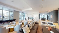 Located at 23 W Chicago Ave in Chicago's vibrant Near North Side neighborhood, this sophisticated amenity lounge is designed to impress with a seamless blend of modern elegance and functional comfort. The space is anchored by a stunning full-height marble feature wall with warm gold veining, housing a sleek linear fireplace and a mounted display screen — creating a dramatic focal point that defines the room's luxurious character.

Light hardwood flooring flows throughout, complementing the crisp white ceilings accented by recessed lighting and a contemporary multi-globe chandelier. Floor-to-ceiling windows wrap the perimeter, flooding the interior with abundant natural light while offering sweeping views of the Chicago skyline and the surrounding streetscape below.

The layout is thoughtfully divided into multiple distinct seating zones. A lounge area features plush sofas, curved barrel chairs, and artful accent tables in mixed metal finishes. A formal dining arrangement with a long dark table and upholstered chairs provides a setting for private gatherings. A media nook, defined by muted slate-toned wall paneling, offers intimate seating in front of a wall-mounted screen. A grand piano adds a refined cultural touch, while a bar-height seating area near the entrance completes the space. Together, these elements create a dynamic, upscale environment ideal for both social and professional use.