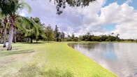 This single-floor property is distinguished primarily by its remarkable outdoor setting, which commands immediate attention. The grounds are generously sized, featuring a large, calm pond that reflects the open sky and surrounding landscape. Well-maintained, vivid green lawns extend along the water's edge, creating a smooth transition between the manicured grounds and the natural waterway. Mature trees of varying species — including palms, weeping willows, and cypress — frame the perimeter, providing natural shade and a layered, verdant backdrop. A wooden split-rail fence is visible in the distance, subtly defining boundaries while preserving the open, pastoral character of the landscape. The overall ambiance is peaceful and park-like, evoking a sense of seclusion and connection to nature. The property's outdoor environment is well-suited for those who appreciate serene waterfront views and lush natural surroundings. The interior spaces of the single-floor structure are noted within the scan, though full interior detail is limited in available imagery. The combination of the expansive grounds, water feature, and mature native vegetation makes the outdoor areas a defining and standout characteristic of this property.