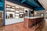 This contemporary coworking space is thoughtfully designed to balance collaborative energy with focused productivity. The open floor plan is anchored by rich hardwood flooring that transitions seamlessly into terracotta brick tile in the kitchen and lounge areas, creating a warm yet dynamic contrast throughout.

The communal kitchen serves as a central social hub, featuring a generously sized island with a marble-topped surface clad in deep red brick tile and lined with sleek black bar stools with red cushioned seats. Behind the island, white cabinetry, open wood and metal shelving, stainless steel appliances, and pendant lighting over a deep navy-painted ceiling create a polished, café-like atmosphere.

The open workstation areas are defined by exposed navy ceilings with visible ductwork, lending an industrial character balanced by warm lighting and curated furnishings. Collaborative seating clusters with rounded tables and colorful accent chairs are interspersed throughout, encouraging spontaneous interaction.

Glass-walled conference and meeting rooms with black steel framing provide visual transparency while offering acoustic privacy, and additional enclosed private offices with warm wood-toned interiors cater to heads-down work. A mezzanine-level overlook with black mesh railings and dramatic hanging pendant lights adds architectural depth and visual intrigue to the overall environment.