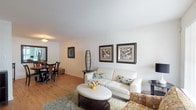 This single-floor residence offers a thoughtfully arranged layout that balances comfort and functionality across its key living spaces. Warm hardwood-style flooring runs consistently throughout, lending a cohesive and inviting feel to the entire unit.

The living room serves as the central gathering space, featuring large windows with vertical blinds that allow natural light to fill the room. Crisp white walls provide a neutral backdrop, and the open flow connects seamlessly to the adjacent dining area, which accommodates a round table with seating for four beneath a ceiling light fixture.

The kitchen is equipped with light wood cabinetry, dark granite-style countertops, and black appliances including a refrigerator and electric range with overhead ventilation. A closeted storage area is conveniently accessible from the kitchen.

The primary bedroom is a well-sized retreat with a large window, hardwood-style flooring, and room for a full seating arrangement alongside the bed. The second bedroom is bright and functional, featuring a window with outdoor views and an open closet with built-in shelving.

The bathroom includes a shower-tub combination, a wood vanity with dark countertop, and hardwood-style flooring, completing this comfortable and practical residence.