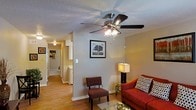 This single-story residence features a welcoming living room as its central gathering space. The room is finished with warm hardwood flooring that extends throughout the hallway, creating a cohesive and continuous flow between areas. A five-blade ceiling fan with an integrated light fixture is mounted to the textured ceiling, providing both air circulation and ambient lighting to the space.

The living area offers comfortable room for seating arrangements, as seen with the red sofa and accent chair, complemented by a glass-top coffee table and a floor lamp that adds a warm, layered lighting option. Neutral beige walls provide a versatile backdrop for décor, with wall art serving as a simple focal point.

A hallway visible from the living area leads deeper into the residence, with recessed lighting overhead guiding the path. White cabinetry is partially visible along the hallway, suggesting proximity to a kitchen or utility area. The overall ambiance of the space is casual and functional, with practical finishes and a straightforward layout that maximizes the usability of the available space.