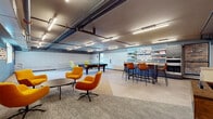 This single-floor amenity and recreation space showcases a contemporary industrial design, characterized by exposed overhead pipes and ductwork, linear strip lighting, and a combination of carpeted and hard-surface flooring throughout. The open layout is divided into distinct activity zones that flow seamlessly into one another, creating a dynamic and social environment.

The lounge area features a cluster of vibrant orange swivel chairs arranged around a low round table, offering a casual and inviting space for conversation or relaxation. Adjacent to this is a bar-height seating section with tan leather stools and tall communal tables, ideal for collaborative work or informal gatherings. Display shelving along the teal-accented wall adds visual interest and a functional organizational element.

A dedicated recreation zone houses a full-size pool table and a foosball table, providing engaging entertainment options. A secondary seating nook near a railing features bold green accent chairs, adding a pop of color to the space. The walls incorporate a mix of teal painted concrete block, warm ochre tiling, and white paneling, contributing to a layered, textured aesthetic. Natural light filters in through select windows, complementing the bright overhead illumination. The space is well-suited for use as a community lounge, amenity room, or shared recreational facility.