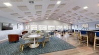 Located at 5001 SW 20th St in Ocala, Florida, this single-floor community gathering space offers a thoughtfully designed interior that balances comfort with functionality. The open-plan layout is anchored by a vaulted ceiling adorned with decorative acoustic panels, which both enhance the room's visual appeal and contribute to a pleasant acoustic environment. Recessed lighting and natural light from a generous row of windows along the exterior wall illuminate the space, offering views of the surrounding outdoor area.

The lounge zone features a large sectional sofa, accent chairs, and pedestal side tables arranged atop a patterned area rug, creating an inviting setting for relaxed conversation. A wall-mounted flat-screen TV adds a media focal point to this area. Adjacent to the lounge, a dining section provides multiple tables paired with modern blue upholstered chairs, well-suited for group gatherings or casual meals.

The kitchenette is equipped with warm wood cabinetry, stone-toned countertops, a stainless steel refrigerator, microwave, and sink — all accented by a striking driftwood and floral wall sculpture that serves as a standout artistic centerpiece. Light wood-look flooring flows seamlessly throughout, tying the entire space together with a cohesive, contemporary aesthetic.