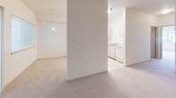 This single-floor residential unit offers a clean, neutral aesthetic with a practical and well-organized layout. The living area is generously sized, featuring soft wall-to-wall carpeting and a sliding glass door dressed with vertical blinds that opens to a view of lush, mature greenery outside, allowing natural light to fill the space.

The kitchen is efficiently designed with light wood cabinetry, ample upper and lower storage, a double sink, and white appliances including a refrigerator, range, and dishwasher. Tile flooring in the kitchen adds durability and ease of maintenance. A pendant light overhead brightens the area, and the space connects openly to the adjacent living and dining areas.

The bedroom is a comfortable, carpeted room with a large window fitted with horizontal blinds, providing both natural light and exterior views. The neutral walls and soft flooring create a calm, restful atmosphere.

The bathroom is well-appointed with an extended vanity countertop, under-sink cabinetry, a wide mirror, and tile flooring. A grab bar is installed for accessibility, and the overall layout is clean and functional.

An additional carpeted room houses the electrical panel and offers flexible utility. Throughout the unit, the consistent use of warm neutral tones, carpet, and tile creates a cohesive and inviting living environment.