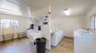 Located at 1515 Broadway in El Cajon, California, this shared laundry facility is a single-floor, well-organized utility space designed to serve multiple users efficiently. The room is laid out to maximize functionality, with a row of commercial-grade top-loading washing machines and dryers lining one wall, providing ample laundry capacity. A dedicated utility sink with a soap dispenser is conveniently positioned for hand-washing or pre-treating garments, accompanied by a centrally placed trash receptacle for easy access. A built-in blue laminate folding counter runs along one side of the room, offering generous surface space for sorting and folding laundry. A wall-mounted wooden garment rod provides a practical area for hanging freshly laundered clothing. The space is illuminated by overhead fluorescent lighting and benefits from natural light through windows fitted with decorative ironwork grilles. Durable stone-pattern tile flooring throughout adds to the room's practicality and ease of maintenance. A bulletin board displaying informational notices and safety signage is mounted near the sink area. A laundry cart is available for residents' convenience. The facility is clean, clearly organized, and designed with shared use in mind, offering all essential amenities in one accessible location.