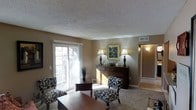This single-floor space offers a comfortable and thoughtfully designed living environment centered around a welcoming living room. The room is finished with warm taupe walls and neutral-toned carpeting that create an inviting atmosphere. A textured ceiling adds architectural character overhead, while white-trimmed sliding glass doors flood the space with abundant natural light and provide access to an outdoor balcony area.

The living room is furnished with a classic arrangement, featuring patterned accent chairs with bold floral motifs, a wooden coffee table, and matching ottomans that lend a cohesive, traditional aesthetic. A dark wood dresser serves as a decorative focal point along the main wall, adorned with matching table lamps, candles, and a framed landscape painting in an ornate gold frame — adding a refined, elegant touch.

A hallway visible from the living area leads further into the space, hinting at additional rooms beyond. The layout feels open and connected, with tile flooring transitioning from carpeted areas near the entryway, adding visual contrast. Overall, the space balances functionality with classic decor, making it a warm and livable environment suitable for everyday comfort.