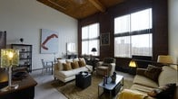 This striking two-floor space showcases a compelling fusion of industrial heritage and contemporary living. The main living area immediately commands attention with its soaring ceilings, rich exposed wooden beam work overhead, and expansive brick accent walls that speak to the building's architectural roots. Large floor-to-ceiling windows dressed with vertical blinds flood the interior with abundant natural light while framing views of the surrounding exterior.

The living area is generously proportioned, accommodating a comfortable seating arrangement that includes a cream upholstered sofa accented with earth-toned throw pillows, classic armchairs, and a substantial coffee table anchored on a textured area rug. A compact dining area sits nearby, maintaining an open and connected feel throughout the space. Decorative touches such as bold wall art and carefully placed lamps contribute to a warm, curated ambiance.

The raw, industrial aesthetic of the brick walls and structural beams is thoughtfully balanced by soft furnishings and refined décor, creating an inviting yet visually dynamic environment. The two-floor layout provides a sense of vertical spaciousness, adding depth and dimension to the overall character of this distinctive and well-appointed space.