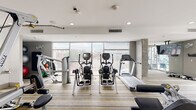 Located at 2110 Lawrence St in Denver, CO, this fitness center offers a thoughtfully designed single-level workout space with a contemporary aesthetic. The facility is defined by its floor-to-ceiling windows along the perimeter, which flood the interior with abundant natural light and frame views of the surrounding Denver skyline and neighborhood streetscape.

The space is laid out to accommodate a diverse range of exercise needs. A solid selection of cardio equipment is prominently featured, including treadmills, elliptical trainers, and stationary bikes, all positioned to take advantage of the expansive window views. Strength training is well represented with multiple cable and selectorized machines from brands including Hoist and Cybex, along with free weights and benches.

Exposed concrete columns contribute an industrial character that complements the clean, modern finishes found throughout. Striped, multi-toned carpeting covers the floor, adding warmth and color to the otherwise neutral palette of white ceilings and light gray walls. Recessed lighting provides consistent illumination throughout the space. A wall-mounted flat-screen television and mirrored wall panels enhance functionality and openness. Glass-paneled doors provide access to an adjacent outdoor area, further connecting the interior to the building's surroundings.