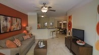 This single-floor residence showcases an open-concept design that seamlessly connects the living, kitchen, and dining areas. The living room serves as a central hub, featuring plush carpeting, a ceiling fan with integrated lighting, and a bold terracotta accent wall that adds warmth and visual interest to the space. A comfortable sofa with decorative throw pillows anchors the seating area, complemented by a wooden coffee table and a media console housing a flat-screen television.

A half-wall breakfast bar with bar stool seating creates a natural transition between the living area and the kitchen, providing both a functional surface and a subtle spatial divider. The kitchen overlooks the adjacent areas, enhancing the open, connected feel of the layout. Framed artwork adorns the walls, contributing to the curated aesthetic.

Beyond the breakfast bar, a dedicated dining area is visible, featuring a chandelier overhead and a full dining table with chairs, making it well-suited for entertaining. Neutral beige tones dominate the walls and flooring throughout, creating a cohesive backdrop that ties the warm accent colors together and gives the overall space a welcoming, modern ambiance.