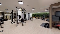 This contemporary fitness center is thoughtfully designed to accommodate a wide range of workout styles and training needs. The space is anchored by a striking living green wall that runs along one side, adding a vibrant, biophilic element that contrasts beautifully with the clean white walls and warm wood accents throughout. Light hardwood-style flooring extends across the entire floor, lending a bright, polished aesthetic underfoot.

Circular flush-mounted ceiling fixtures provide consistent, even illumination throughout the space, creating a well-lit environment conducive to focused training. Equipment is organized to maximize the open floor plan, with dedicated zones for cardio, strength, and functional training. Cardio offerings include Peloton stationary bikes and treadmills, while strength training is supported by a Life Fitness cable and functional trainer system, a lat pulldown machine, a squat rack, and an extensive dumbbell rack. Kettlebells, parallette bars, a stability ball, and a stall bar round out the functional fitness options. Full-length mirrors along one wall enhance spatial depth and allow for form monitoring. A separate adjoining area with glass partitions provides additional utility space, completing this comprehensive and modern fitness facility.