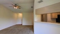 This single-floor residence offers a straightforward and functional layout designed for comfortable everyday living. The main living area is spacious and open, featuring wall-to-wall carpeting that adds warmth underfoot. A ceiling fan with a built-in light fixture is centrally positioned overhead, providing both circulation and ambient lighting. The textured ceiling adds a classic residential touch throughout the space.

A defining architectural feature is the pass-through window cutout that connects the kitchen area to the living room, creating a sense of openness between the two spaces while maintaining a defined separation. The kitchen area is partially visible through this opening, hinting at a practical galley-style layout with louvered cabinet doors.

Neutral, light-toned walls contribute to a bright and airy atmosphere, while a paneled entry door provides a clean and welcoming front entrance. HVAC vents are visible in the ceiling, ensuring climate control throughout the home. The overall design is practical and unfussy, making the space easy to personalize and arrange to suit a variety of lifestyles. The single-floor layout ensures convenient, step-free access to all areas of the home.