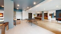 Located at 2702 Pelham Rd in Greenville, SC, this single-floor communal lounge is a thoughtfully designed gathering space that blends modern aesthetics with social functionality. The open-plan layout is anchored by a signature teal-painted ceiling that contrasts elegantly with crisp white walls and light-toned tile flooring, which transitions into a warm herringbone wood pattern near the café and gaming area.

The main lounge zone features a generous sectional sofa in cognac leather, complemented by rounded dark wood coffee tables and a curated gallery wall showcasing an eclectic mix of framed artwork. A large flat-screen TV is mounted within a built-in shelving unit, creating a dedicated media corner ideal for group viewing. Adjacent accent chairs and side tables provide additional seating for conversation or relaxation.

Structural columns punctuate the open floor plan while a ping-pong table serves as a central recreational focal point, reinforcing the space's social character. Toward the back, a stylish bar and café nook features pendant lighting, counter seating with green stools, and rich red accent walls, offering a vibrant contrast to the lounge's neutral palette. Floor-to-ceiling windows and glass doors flood the space with natural light and provide views of an outdoor terrace, seamlessly connecting interior and exterior environments.