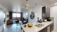 This single-floor residence showcases an open-concept layout that seamlessly connects the kitchen, dining, and living areas into one cohesive living space. The kitchen is appointed with light quartz countertops, a double stainless steel sink, and a tile backsplash, complemented by dark cabinetry that adds contrast and depth. Pendant lights hang from the ceiling, providing focused illumination over the kitchen island, which serves as a natural dividing point between the kitchen and the rest of the living space.

The dining area features a round table with mixed seating, positioned beneath additional pendant lighting and near a decorative sunburst mirror that serves as a striking focal point on the white wall. Track lighting on the ceiling offers flexible illumination throughout. The living area beyond is generously proportioned, with a ceiling fan, hardwood-style flooring, and large windows and a glass sliding door that lead to an outdoor balcony, allowing natural light to fill the interior and providing views of the surrounding area. The overall ambiance is modern and well-appointed.