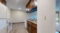 This single-floor residential unit offers a practical and functional layout spread across several well-defined living areas. The kitchen is equipped with dark wood cabinetry providing ample upper and lower storage, a double stainless steel sink with granite-style countertops, a white electric range with overhead ventilation, and a full-size refrigerator. Light wood-style vinyl flooring runs throughout the kitchen and dining area, lending a clean, easy-to-maintain finish. The living room is generously proportioned, featuring neutral carpet, baseboard heating, a wall-mounted air conditioning unit, and a sliding glass door that opens to a private balcony, allowing for natural light and outdoor views. The bedrooms are carpeted and bright, each offering sliding closet doors with built-in shelving for organized storage. Natural light filters in through windows with views of surrounding greenery. The bathroom includes a full-size soaking tub and shower combo with a grab bar, finished in clean white surrounds. Throughout the unit, consistent wood-tone trim, recessed lighting in the kitchen, and neutral wall colors contribute to a cohesive and understated aesthetic.