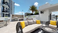 This rooftop terrace is a well-appointed outdoor amenity space designed for both relaxation and entertainment. The terrace features a clean, contemporary aesthetic defined by crisp white perimeter walls, light-toned porcelain tile flooring, and an open-sky setting that invites abundant natural light throughout the day. At the heart of the space, a generously sized wicker sofa set with neutral cushions and bold yellow accent pillows provides a comfortable lounge area, complemented by a stylish round fire pit nearby. An outdoor dining table with chairs offers a dedicated space for al fresco meals, while a built-in BBQ grill station with a decorative mosaic tile accent wall serves as a functional and visually striking cooking area. Lush tropical palm trees and greenery planted along the perimeter railing add a vibrant, resort-like atmosphere to the setting. Wall-mounted industrial fans ensure comfort in warm weather. The open views of the surrounding cityscape and sky further enhance the ambiance, making this rooftop terrace an inviting destination for gatherings, outdoor dining, and leisurely relaxation.