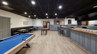 Located at 99 Gold Star Highway in Groton, CT, this single-floor lounge offers a generously sized, open interior designed for leisure and social gathering. The space is anchored by a large central bar with a warm wood-topped counter, bar stool seating on multiple sides, and a brick accent backdrop — creating a natural hub for the room. Recessed lighting set against a dark drop ceiling gives the lounge a sleek, contemporary ambiance, while light wood-style flooring throughout lends a clean, welcoming feel.

The recreational area features a full-size pool table with built-in cabinetry and stone-top counters along the wall, as well as a foosball table centrally positioned for easy access. A dedicated booth seating section lines one wall, furnished with dark upholstered booths and café-style tables, offering a comfortable space for dining or conversation. Decorative frosted glass panels featuring nautical imagery — including bridges and sailing vessels — add a distinctive, locally inspired character to the space.

Framed nautical artwork adorns the walls, reinforcing the maritime theme befitting the Groton, CT location. The layout flows openly from the bar to the game area and seating zones, making this lounge well-suited for both casual visits and larger social events.