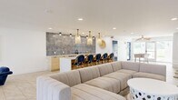 Located at 2045 East Bay Dr in Largo, Florida, this single-floor community space offers a well-appointed, open-plan layout that seamlessly combines a fully equipped kitchen with a spacious living and lounge area. The interior design embraces a coastal aesthetic throughout, with a neutral palette of whites, warm woods, and navy blue accents that create a relaxed yet refined atmosphere.

The kitchen is a standout feature, centered around a large white quartz island with seating for multiple guests. Light wood cabinetry lines the back wall, complemented by a striking floor-to-ceiling textured tile backsplash in muted blue and grey tones. Woven rattan pendant lights hang above the island, reinforcing the beachy ambiance. Stainless steel appliances, including a dishwasher and beverage refrigerator, add modern functionality.

The living area is generously proportioned, anchored by a large wraparound sectional sofa in neutral upholstery and a sculptural round coffee table. A wall-mounted large-screen television serves as a focal point, flanked by decorative surfboards that nod to the Florida coastal lifestyle. A separate seating nook features a curved navy velvet banquette with round bistro tables on brass pedestals.

Large windows and glass doors flood the space with natural light and provide views of the surrounding outdoor pool area, enhancing the open, airy feel of this thoughtfully designed gathering space.