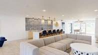 Located at 2045 East Bay Dr in Largo, Florida, this single-floor community space offers a well-appointed, open-plan layout that seamlessly combines a fully equipped kitchen with a spacious living and lounge area. The interior design embraces a coastal aesthetic throughout, with a neutral palette of whites, warm woods, and navy blue accents that create a relaxed yet refined atmosphere.

The kitchen is a standout feature, centered around a large white quartz island with seating for multiple guests. Light wood cabinetry lines the back wall, complemented by a striking floor-to-ceiling textured tile backsplash in muted blue and grey tones. Woven rattan pendant lights hang above the island, reinforcing the beachy ambiance. Stainless steel appliances, including a dishwasher and beverage refrigerator, add modern functionality.

The living area is generously proportioned, anchored by a large wraparound sectional sofa in neutral upholstery and a sculptural round coffee table. A wall-mounted large-screen television serves as a focal point, flanked by decorative surfboards that nod to the Florida coastal lifestyle. A separate seating nook features a curved navy velvet banquette with round bistro tables on brass pedestals.

Large windows and glass doors flood the space with natural light and provide views of the surrounding outdoor pool area, enhancing the open, airy feel of this thoughtfully designed gathering space.
