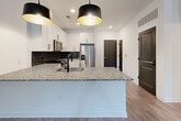 Located at 397 N Central Ave in Hapeville, GA, this thoughtfully designed one-bedroom, one-bathroom residence offers a modern aesthetic with quality finishes across a single floor. The open-concept kitchen and living area serves as the heart of the home, anchored by a large granite-topped island with an undermount sink and bold matte black pendant lighting overhead. The kitchen features white shaker cabinetry, a dark subway tile backsplash, and a full suite of stainless steel appliances, including a French door refrigerator, range, over-the-range microwave, and dishwasher. Wood-look flooring runs throughout the main living areas, complementing the crisp white walls and clean architectural lines. The living room receives natural light through a front-facing door flanked by windows, with a ceiling fan adding both function and style. The bedroom is carpeted for comfort, includes a ceiling fan, and offers a window with outdoor views. It connects directly to the bathroom, which features a double-sink granite vanity with white cabinetry and matte black fixtures, plus access to a dedicated closet. The unit presents a cohesive, contemporary design with a neutral palette and modern hardware details throughout.