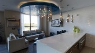 This contemporary single-floor space makes a bold design statement through its carefully curated architectural and decorative elements. The focal point of the room is a dramatic oval tray ceiling finished in a deep teal, metallic tone, which anchors the open-plan layout and adds a sense of depth and sophistication. Suspended from this feature ceiling are sculptural pendant lights with a crinkled, chrome finish that cast a warm, ambient glow throughout the space.

The lounge area is furnished with a comfortable gray sofa accented with teal throw pillows, positioned near a large window that allows natural light to pour in and offers views of the surrounding greenery. A wall-mounted flat-screen television is displayed against a textured, three-dimensional white accent wall that adds visual interest and contrast to the room.

Adjacent to the lounge is a counter seating area featuring a sleek white surface paired with dark, modern bar-style chairs. Additional sculptural pendant lights line the wall above, maintaining a cohesive design language throughout. The overall ambiance is polished and contemporary, blending metallic accents, textural contrasts, and thoughtful lighting for an upscale, inviting atmosphere.