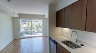 This single-floor space showcases a contemporary open-concept design that seamlessly connects the kitchen and living areas. The kitchen is appointed with rich, dark wood cabinetry featuring under-cabinet lighting, light-toned countertops, and a stainless steel sink with a modern arc faucet. A stainless steel dishwasher adds a practical, polished touch to the culinary space.

The living area extends naturally from the kitchen, finished with warm hardwood-style flooring that runs throughout, contributing to a cohesive and inviting atmosphere. Clean white walls and recessed lighting keep the interior feeling bright and well-illuminated.

A standout feature of this space is the expansive floor-to-ceiling sliding glass door that opens to a balcony, flooding the interior with abundant natural light. The outlook reveals lush tree canopies and a broader urban landscape, offering a refreshing connection to the outdoors. The overall design balances modern finishes with functional layout, creating a clean, contemporary living environment well-suited for everyday comfort.