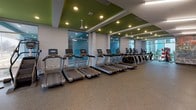 This single-floor fitness center is thoughtfully designed to accommodate a full range of workout activities within a spacious, open layout. The facility is divided into distinct functional zones, each optimized for a specific type of training. The cardio area runs along a wall of large floor-to-ceiling windows, allowing abundant natural light to fill the space while providing outdoor views. A row of modern treadmills with individual screens lines this windowed wall, accompanied by ellipticals and other aerobic machines. Ceiling fans mounted above the cardio zone enhance comfort during workouts.

The strength training area features a comprehensive selection of Hoist cable machines and selectorized equipment arranged along one side of the facility. A dedicated free weights zone offers a full rack of dumbbells, adjustable benches, medicine balls, and additional weight training accessories, set against soft green accent walls with mirrored surfaces. An open floor area provides space for functional training.

Motivational details are woven throughout the design, including a bold graffiti-style mural on an exposed brick accent wall and large typographic "HYDRATE" signage near the water fountain station. The durable speckled rubber flooring, recessed lighting, and green ceiling panels create a cohesive, energetic aesthetic throughout the entire facility.