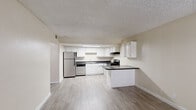 This single-floor unit is organized around a functional, open layout that connects the kitchen and living areas seamlessly. The kitchen is a standout feature, equipped with white raised-panel cabinetry offering generous storage across both upper and lower cabinets. Dark speckled countertops provide a contrast against the white cabinetry, and the space is outfitted with stainless steel appliances, including a refrigerator, electric range, and dishwasher. An under-cabinet range hood and flush-mount fluorescent lighting keep the workspace well-lit. A peninsula with a dark countertop extends from the kitchen, defining the boundary between the cooking and living areas.

The living area benefits from natural light entering through a large window fitted with vertical blinds, creating a bright and open atmosphere. Light wood-style flooring runs throughout the kitchen and living spaces, adding a clean, cohesive aesthetic. The bedroom features plush carpeting and its own window with vertical blinds, offering a quieter, comfortable retreat.

The bathroom includes a dual-sink vanity topped with dark granite-style countertops, white cabinetry below, paired mirrors, and a well-lit vanity bar above. Light wood-style flooring continues here, maintaining a consistent look throughout. Textured popcorn ceilings are present throughout the unit.