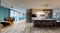 This thoughtfully designed clubhouse offers a dynamic, single-floor community space that balances social activity with comfortable retreat. Throughout the space, wood-look tile flooring provides a warm, cohesive foundation that ties together each distinct zone.

The main lounge area serves as a central gathering point, anchored by a large wall-mounted flat-screen TV and a sleek linear fireplace set against a rich wood-paneled accent wall. Generous seating — including modular sofas, accent chairs, and ottomans — is arranged to encourage conversation, while floor-to-ceiling windows flood the room with natural light and offer views of an outdoor pool area.

Adjacent to the lounge, a game zone features a full-size pool table and additional game tables, ideal for casual recreation. The communal bar area is a standout feature, showcasing a long countertop with a light stone surface, bar seating, and a striking rustic wood beam pendant light fixture with Edison-style bulbs hanging overhead. A mounted TV and counter-level sink complete the functional setup.

A dedicated co-working area provides semi-private booths with teal upholstered seating, offering a quieter environment for focused work. The reception area near the entrance is finished with a reclaimed wood-accented desk and additional lounge seating. Stone accent columns and slatted wood dividers add architectural texture throughout.
