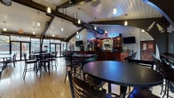 This bar and lounge is a well-appointed single-floor venue designed to accommodate a lively social atmosphere. The space is defined by its striking vaulted ceiling with exposed dark structural beams and white tongue-and-groove paneling overhead, giving the interior an open, airy quality. Industrial-style ductwork runs along the roofline, adding a distinct character to the overall aesthetic. Pendant lighting is suspended throughout, casting a warm, inviting glow across the room.

The centerpiece of the space is a long, fully stocked bar finished in rich wood tones, lined with bar stools and backed by an extensive selection of spirits and beer taps. Multiple wall-mounted flat-screen televisions are positioned throughout for optimal visibility from all seating areas. Neon signage and branded décor add a casual, energetic ambiance.

The main floor is finished with light hardwood flooring and is furnished with a mix of high-top pub tables and chairs arranged across several seating zones. Large windows and glass doors along the perimeter flood the space with natural light and offer views of an outdoor deck and surrounding trees. A dartboard and other recreational features are also present, enhancing the entertainment offerings of this well-rounded venue.