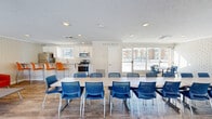 This single-floor community space is thoughtfully designed to serve as a versatile gathering hub, combining a kitchen, dining room, and lounge area within a cohesive, well-appointed layout. Throughout the space, white-painted brick walls lend a clean, classic character, while wood-style flooring adds warmth underfoot.

The fully equipped kitchen features stainless steel appliances — including a gas range, microwave, and refrigerator — complemented by white shaker cabinetry, a tile backsplash, and granite countertops. A peninsula with bar-height seating accommodates casual dining, with vibrant orange bar stools adding a bold pop of color.

The dining area is generously furnished with long white tables and modern blue mesh chairs, offering ample seating for group gatherings or events. Large sliding glass doors and expansive windows flood the dining space with natural light and provide open views of the surrounding exterior. An additional smaller dining table near the glass doors offers a more intimate setting.

The lounge area is anchored by plush red accent chairs arranged around a low coffee table, creating a comfortable and inviting space for relaxed conversation. Large windows continue to draw in natural light, reinforcing the bright, airy ambiance that defines the space. Recessed lighting and textured ceilings complete the well-maintained, functional interior.