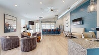 Located at Authentix Apartments in Greeley, Colorado, this single-floor community clubhouse delivers a contemporary, open-concept design with a welcoming and functional atmosphere. Upon entry, guests are greeted by a striking blue reception counter with granite-style surfaces and pendant lighting, establishing a polished first impression. Exposed silver ductwork runs overhead, adding an industrial touch that contrasts beautifully with the bright white walls and light wood-look flooring throughout.

The main gathering area features a comfortable lounge seating arrangement centered around a low coffee table, with a wall-mounted flat-screen TV and ample natural light streaming through large windows. Adjacent to the lounge, a dedicated workspace area with computer stations provides a productive environment for residents.

A standout architectural feature is the full-width garage-style door that opens directly to an outdoor patio and pool area, seamlessly blending indoor and outdoor living. Nearby, a well-equipped kitchenette with white cabinetry, granite countertops, stainless steel appliances, and a built-in microwave serves residents with everyday convenience.

A blue shiplap accent wall adorned with longhorn décor anchors a bar-height counter with leather-accented stools, ideal for casual dining or socializing. Recessed lighting and a ceiling fan complete the bright, airy ambiance of this thoughtfully designed community space.