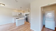 Located along S Burnsfield Ave in Sioux Falls, South Dakota, this single-floor unit offers a clean, contemporary design with a cohesive neutral color palette and light wood-style flooring that runs consistently throughout the space.

The kitchen serves as a central highlight, outfitted with crisp white shaker-style cabinetry featuring matte black hardware, light countertops, and a full suite of stainless steel appliances including a refrigerator, electric range, over-the-range microwave, and dishwasher. A center island provides additional prep and counter space, while a pendant light overhead adds a functional decorative touch. Recessed lighting and vaulted ceiling lines in portions of the kitchen contribute to an open, airy feel.

The floor plan, as illustrated by the overhead model, reveals a well-organized layout with distinct zones for living, cooking, and utility purposes. A dedicated laundry area includes a stacked washer and dryer unit alongside generous wire shelving for storage.

The bathroom presents a tidy, modern aesthetic with a white vanity, dark-veined countertop, matte black fixtures and accessories, a wall mirror, and hardwood-style flooring that continues from the main living areas. The overall ambiance of the space is bright, functional, and move-in ready.
