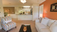 This single-floor space showcases an open-concept layout that connects the kitchen and living area into one cohesive, functional environment. The kitchen is outfitted with white cabinetry, a stainless steel refrigerator, white appliances including a built-in microwave and stove, and granite-style countertops. A peninsula counter extends outward, creating a natural divider between the kitchen and living space while also serving as a casual dining or prep area. Overhead, recessed lighting and under-cabinet fixtures keep the kitchen well-illuminated.

The living area features warm beige carpeting and is accented by a bold terracotta-orange wall that adds visual depth and character to the space. A ceiling fan with a light fixture is centrally mounted, providing both air circulation and ambient lighting. The layout accommodates comfortable seating arrangements centered around a dark coffee table, with ample room to move freely. A set of louvered closet doors along one wall provides generous storage. The overall ambiance is warm and inviting, with a cohesive neutral palette punctuated by vibrant accent tones.