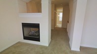 This two-story residence offers a welcoming and functional layout designed for comfortable living. The main living area is anchored by a classic fireplace framed by a white mantel and set into a structural column, adding both architectural interest and a cozy focal point to the room. Soft, wall-to-wall carpeting in a warm beige tone runs throughout, contributing to the home's understated and inviting ambiance.

The neutral color palette — consisting of warm whites and soft beige tones on the walls and trim — creates a cohesive and light-filled atmosphere across the space. White painted trim and column detailing add a touch of traditional character to the interior. The open sightlines from the living area extend down a hallway toward what appears to be additional rooms, suggesting a well-connected and spacious floor plan.

A glimpse of natural light visible at the end of the corridor hints at generously placed windows throughout the home. The second floor adds additional living or sleeping space, expanding the overall functionality of the residence. The clean, move-in-ready condition of the interior makes it easy to envision how each space can be personalized and furnished to suit a variety of lifestyles and needs.