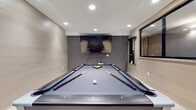 This contemporary entertainment space is thoughtfully designed to offer a dynamic and inviting recreational experience. At the heart of the room sits a full-size pool table with a sleek grey felt surface and dark frame, positioned beneath recessed ceiling lighting that illuminates the play area with clarity and precision. A large wall-mounted flat-screen TV anchors the focal wall, which is clad in a distinctive textured grey tile accent wall, adding depth and visual interest to the space. The remaining walls are finished in light, horizontal wood-plank paneling, lending a warm, modern aesthetic throughout. Light-toned hardwood flooring complements the overall palette and ties the design together cohesively. A standout architectural feature is the sunken lounge alcove, set slightly below floor level and carpeted in grey, where oversized bean bag chairs are arranged for casual, comfortable seating. This nook is framed by the same textured accent wall, creating an intimate retreat within the larger room. A large pass-through window with black framing connects to an adjacent area, suggesting a service or bar counter nearby. The space is accessed through glass-paneled doors, and recessed lighting throughout ensures a well-lit yet relaxed ambiance suited for both active recreation and leisure.