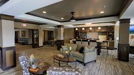 This single-floor community space exudes a warm and sophisticated ambiance, blending modern design with inviting comfort. The open-plan layout seamlessly connects the main lounge area to the reception and kitchenette, creating a cohesive and functional environment. The lounge is anchored by plush, neutral-toned sofas and armchairs arranged around a rustic wooden coffee table, offering a relaxed setting for socializing or unwinding. A ceiling fan with pendant-style lighting adds a refined touch overhead, while recessed lighting throughout ensures a bright and welcoming atmosphere. Structural columns with dark accents frame the space elegantly, complementing the rich, dark wood tones found in the cabinetry and reception counter. The kitchenette area features dark wood cabinetry, a mosaic tile backsplash, and pendant lighting, lending a polished, café-like feel. The reception desk is neatly positioned nearby, providing a clear point of welcome for guests and residents. Patterned carpeting in the lounge area adds texture and visual interest, while wood-look flooring in adjacent areas ties the overall design together. The thoughtful layout and curated décor make this a versatile and stylish communal hub.