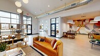 This expansive single-floor amenity space offers a generously sized, open-concept layout designed for both socializing and relaxation. The interior is defined by soaring ceilings with exposed industrial-style ductwork, light hardwood-style flooring, and an abundance of natural light streaming through large black-framed windows and clerestory windows that line the upper walls of the game room area.

The main lounge features a tufted caramel leather sofa anchored by a striped area rug, complemented by a freestanding open-shelf display unit and globe pendant lighting. Adjacent to the lounge, a dining and casual seating area showcases rich colorblock wall art in teal, yellow, and gold tones, with leather banquette seating, round marble-top tables with gold pedestal bases, and woven accent chairs.

The kitchenette is outfitted with a granite-topped island bar, subway tile backsplash, slate-blue cabinetry, and warm brass pendant lights — ideal for entertaining. A dedicated game room boasts soaring double-height ceilings, floor-to-ceiling windows with sweeping outdoor views, a ping-pong table, and vibrant wall art. Glass-panel French doors connect the distinct zones while maintaining an open, airy flow throughout the entire space.