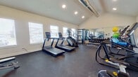 This fitness center offers a well-organized and fully equipped workout space designed to support a wide range of exercise routines. The room features vaulted ceilings that create an open, airy atmosphere, enhanced by recessed lighting and multiple windows fitted with blinds that allow natural light to fill the space throughout the day. Exposed ductwork runs along the upper portion of the ceiling, adding an industrial accent to the otherwise clean, neutral interior.

The dark rubber flooring spans the entire area, providing a durable and slip-resistant surface suitable for all types of exercise. Cardio equipment is well-represented, with multiple treadmills, an elliptical trainer, a stair climber, and a stationary spin bike positioned throughout the room. A multi-station cable machine offers versatile strength training options, while a dedicated free weight area includes a full dumbbell rack and a flat bench for targeted resistance work.

A large wall-mounted mirror on one side visually expands the space and allows users to monitor their form during workouts. Additional accessories such as stability balls and medicine balls on a storage rack add further variety to available training options. Glass entry doors provide a welcoming entry point, and the overall layout ensures comfortable spacing between equipment for safe, efficient use.