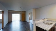 This single-floor residence offers an open-concept design that seamlessly connects the kitchen and living areas. The kitchen features granite countertops with a light speckled finish and dark cabinetry, providing a clean, modern aesthetic. The countertop extends as a partial divider, naturally separating the kitchen from the main living space without disrupting the open feel.

The living area is finished with dark hardwood-style flooring that runs throughout, adding warmth and visual continuity. A large sliding glass door with vertical blinds floods the space with natural light while providing access to an outdoor area. The neutral grey walls complement the flooring and create a calm, cohesive palette throughout.

A carpeted room is visible through an interior doorway, likely serving as a bedroom, and benefits from its own natural light source. A second interior door with a warm wood finish leads to what appears to be a utility or storage area. The smooth white ceilings and clean wall lines contribute to the overall sense of an organized, functional layout well-suited for everyday living.