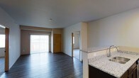 This single-floor residence offers an open-concept design that seamlessly connects the kitchen and living areas. The kitchen features granite countertops with a light speckled finish and dark cabinetry, providing a clean, modern aesthetic. The countertop extends as a partial divider, naturally separating the kitchen from the main living space without disrupting the open feel.

The living area is finished with dark hardwood-style flooring that runs throughout, adding warmth and visual continuity. A large sliding glass door with vertical blinds floods the space with natural light while providing access to an outdoor area. The neutral grey walls complement the flooring and create a calm, cohesive palette throughout.

A carpeted room is visible through an interior doorway, likely serving as a bedroom, and benefits from its own natural light source. A second interior door with a warm wood finish leads to what appears to be a utility or storage area. The smooth white ceilings and clean wall lines contribute to the overall sense of an organized, functional layout well-suited for everyday living.