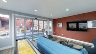 This single-floor communal lounge and entertainment space is thoughtfully designed to blend indoor comfort with outdoor accessibility. The interior is defined by bold terracotta-red accent walls that contrast with crisp white ceilings and light hardwood-style flooring, creating a vibrant yet inviting atmosphere. Recessed lighting illuminates the space evenly, enhancing its modern aesthetic.

A centerpiece of the room is a full-length shuffleboard table, offering a standout recreational feature for gatherings. Adjacent to it, a generously sized blue sectional sofa anchors a relaxed seating area, complemented by contemporary accent chairs and a glass-top coffee table — ideal for casual socializing or media viewing. A wall-mounted flat-screen television is prominently positioned on the accent wall, making this area well-suited for entertainment.

Large multi-panel glass folding doors span one full wall, flooding the interior with natural light and providing seamless access to an outdoor patio area. The patio, visible through the glass, features additional seating arrangements beneath ceiling fans, extending the usable social space outdoors. The overall design emphasizes openness, flow, and a lively communal character throughout.