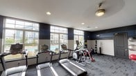 This fitness center is designed to provide a functional and visually appealing workout environment. The space is characterized by a bold two-tone color scheme, with deep navy accent walls contrasting against crisp white upper walls and ceiling, lending a modern and energetic atmosphere. Recessed lighting, complemented by a ceiling fan, ensures the space is well-lit and ventilated throughout.

Large, floor-to-ceiling windows span one wall, flooding the room with natural light and offering direct views of an outdoor pool and surrounding residential buildings, creating a bright and motivating setting for exercise. The windows' multi-pane design adds an architectural element to the space.

The fitness area is equipped with a focused selection of cardio equipment, including multiple treadmills and an elliptical trainer, all positioned to take advantage of the poolside views. A stationary bike and additional cardio equipment round out the selection. Along the opposite wall, a dedicated free-weight station features a rack of medicine balls, providing options for strength and functional training.

Durable, low-pile carpet in a neutral grey tone covers the floor, offering comfort and practicality underfoot. The overall layout is open and efficient, making effective use of the available space while maintaining an uncluttered, accessible environment for users.