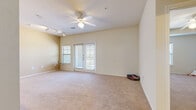 This single-floor residence offers a practical and comfortable layout with a variety of well-designed living spaces. The main living area is generously sized, finished with plush carpeting, and illuminated by a ceiling fan with integrated lighting. Track lighting along one wall provides additional ambiance, while a set of French doors flanked by windows invites natural light and opens to the outdoors. A secondary carpeted room, also featuring a ceiling fan and dual windows with views of surrounding greenery, serves well as a bedroom or flexible living space. The kitchen is thoughtfully appointed with warm wood cabinetry, granite-style countertops, stainless steel appliances including a refrigerator and range, and durable hardwood-style flooring. A set of bifold doors reveals a dedicated laundry closet directly off the kitchen. The primary bathroom is elongated and features a vanity with wood cabinetry, a large mirror, a separate glass-enclosed shower, and hardwood-style flooring. A second full bathroom offers a shower-tub combination and matching vanity. A spacious walk-in closet with built-in wire shelving provides ample storage. Warm-toned hardwood-style flooring runs throughout the hallways, connecting each area seamlessly.